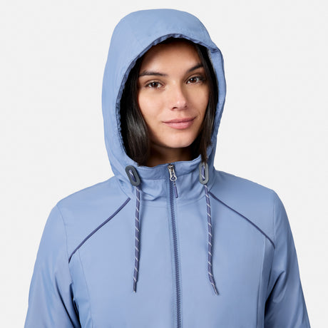 A woman in a light blue Free Country Women’s Anorak Windshear Jacket, featuring recycled polyester, a zipper, and drawstrings, smiles softly at the camera. A plain white background highlights the eco-friendly outerwear.