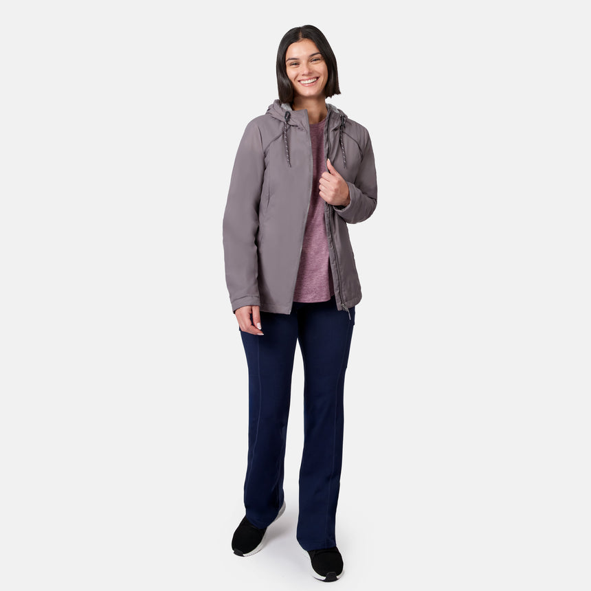 A woman with shoulder-length dark hair smiles in a Free Country Women’s Anorak Windshear Jacket made of gray recycled polyester, paired with a mauve shirt, navy pants, and black shoes against a plain white background.