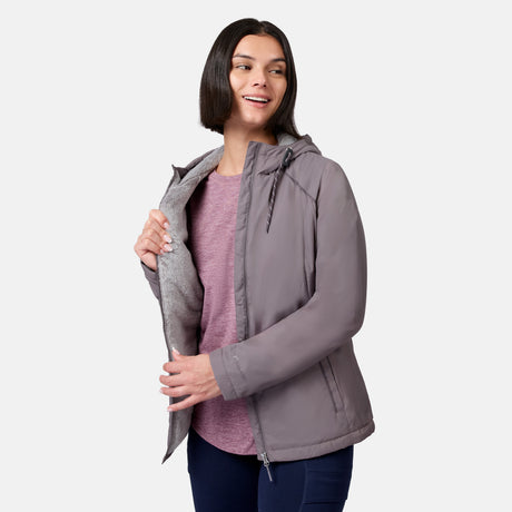 A woman with straight dark hair smiles as she opens her Free Country Women’s Anorak Windshear Jacket to reveal its gray lining. She wears a pink shirt and stands against a plain light background.