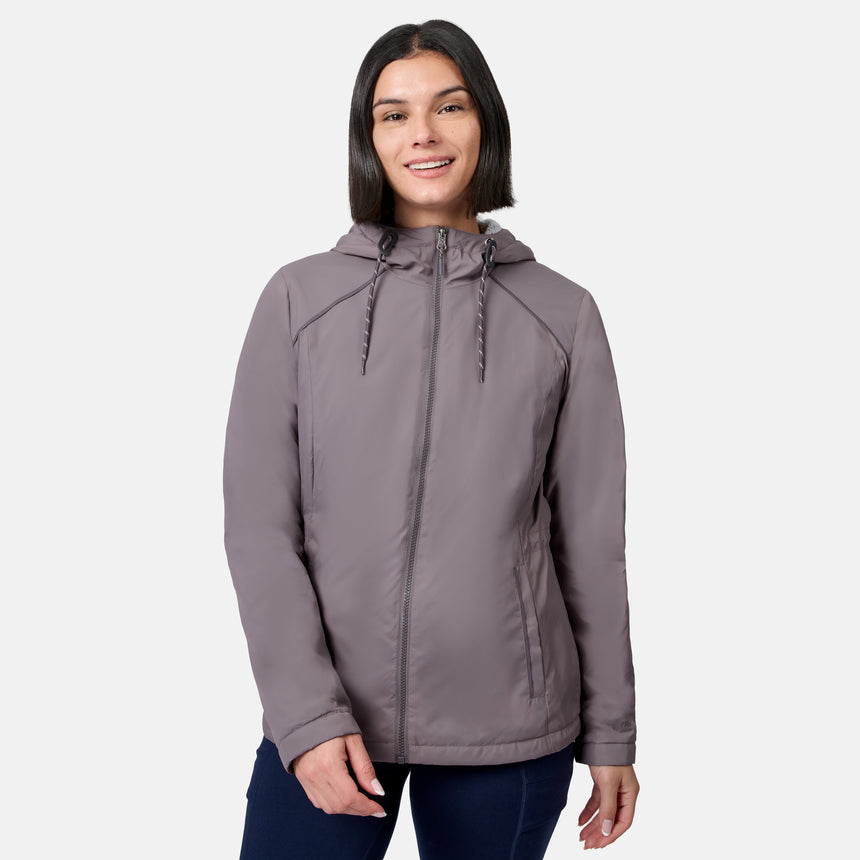 A woman with dark hair, wearing the Free Country Women’s Anorak Windshear Jacket in Zinc and dark pants, smiles while standing against a plain white background.