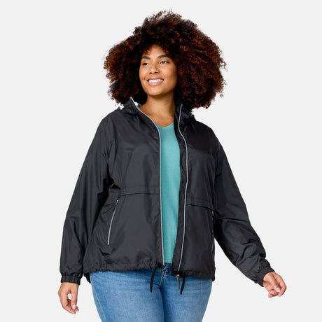 A woman smiles with one hand slightly raised, wearing the Clearance Women's Plus Size Outland Windshear Jacket in black over a teal shirt and blue jeans against a light gray background.