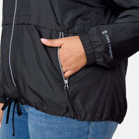 A person wears the Women's Plus Size Outland Windshear Jacket by Clearance—a black, lightweight, water-resistant jacket with a drawstring hem and branding on the sleeve. Their right hand is in the pocket, and they are paired with blue jeans.
