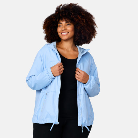 A woman with curly hair smiles in the Women's Plus Size Outland Windshear Jacket by Clearance, a lightweight light blue hooded jacket worn over a black top, standing against a plain light background.
