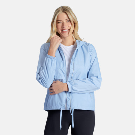 A woman with long blonde hair smiles in the Clearance Women’s Outland Windshear Jacket, a lightweight light blue zip-up, styled over a white shirt and black leggings against a plain white background.