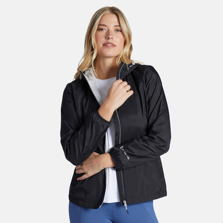 A blonde woman wears the Clearance Women's Trek Mate Windshear Jacket in black over a white shirt and blue pants, standing against a plain light background. She holds the jacket collar with one hand and rests her other hand on her hip.
