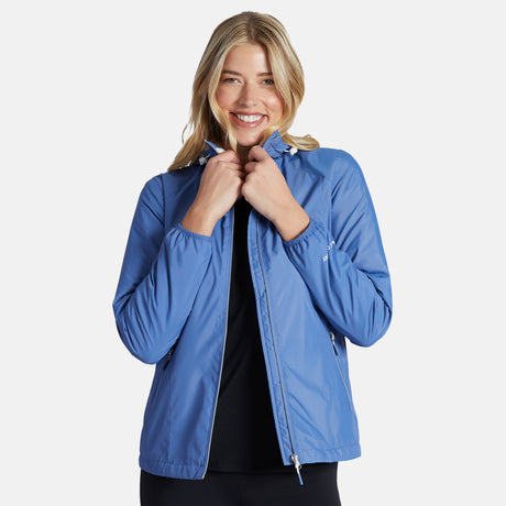 A smiling blonde woman stands against a white background, adjusting the collar of her blue Women's Trek Mate Windshear Jacket by Clearance over a black top.
