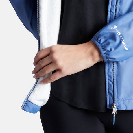 A person is unzipping a Clearance Women's Trek Mate Windshear Jacket in blue, showing a black shirt underneath. Only the torso and hands are visible; the jacket features a white fleece lining and is water-resistant.