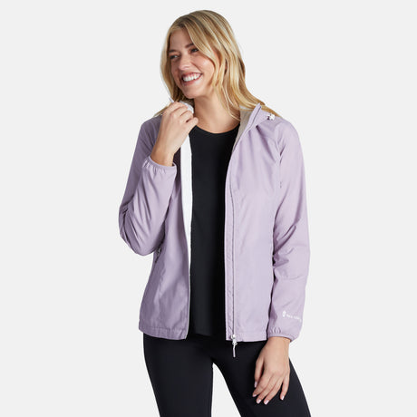 A smiling woman with long blonde hair wears the Clearance Women's Trek Mate Windshear Jacket in light purple over a black shirt and black pants, standing against a plain white background.