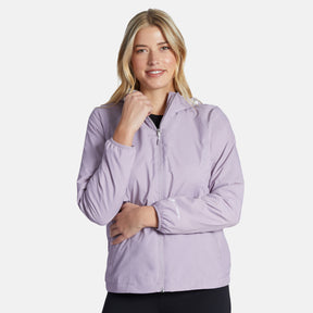A woman with blonde hair models the Clearance Women's Trek Mate Windshear Jacket in light purple, standing against a plain white background, smiling with one hand on her chin and the other arm crossed.