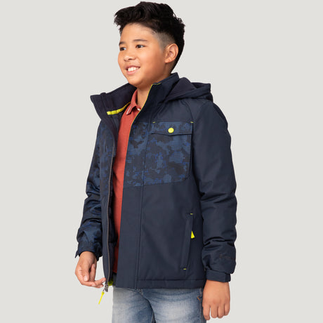 A smiling boy wears the Free Country Boys' Multi Rip Stop Boarder Jacket, a water-resistant kids jacket in navy blue with patterns, over a red polo shirt and light blue jeans, standing against a light gray background.