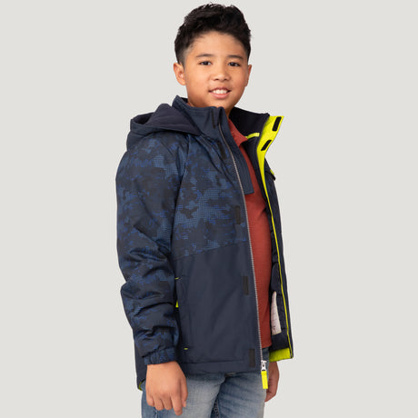 A boy with short dark hair, wearing the Free Country Boys' Multi Rip Stop Boarder Jacket in navy blue over a red shirt and light blue jeans, stands smiling against a plain light background.