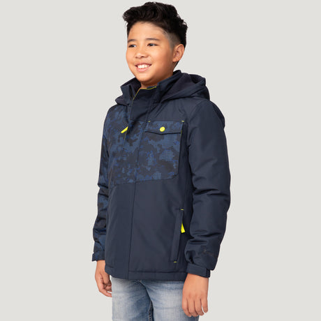 A boy with short, dark hair is smiling while wearing the Free Country Boys' Multi Rip Stop Boarder Jacket in navy blue with yellow accents and a patterned chest, paired with light blue jeans against a plain light gray background.