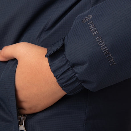A close-up of a child wearing the Free Country Boys' Multi Rip Stop Boarder Jacket in navy blue, featuring water-resistant fabric, a hand in the pocket, and an embroidered Free Country logo on the sleeve near the wrist.