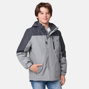 [River is wearing size M.] Young boy wearing the Free Country Boys' 3-in-1 Systems Jacket in a light grey with a hood and subtle chest pattern, smiling against a plain background.