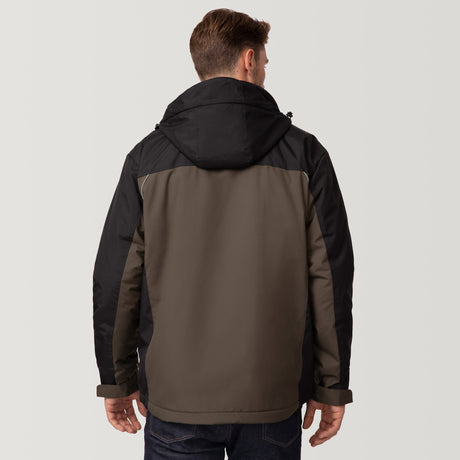 A man faces away from the camera, wearing the Clearance Men's FreeCycle® Jack Frost 3-in-1 Systems Jacket in black and olive green with dark jeans. The jacket is made from recycled REPREVE materials. He stands against a plain light gray background.