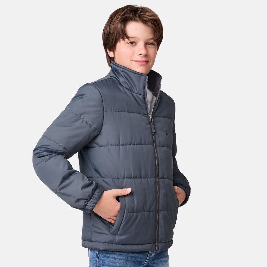 [River is wearing size M.] Young boy wearing the Free Country Boys' 3-in-1 Systems Jacket in a light grey with a hood and subtle chest pattern, smiling against a plain background.