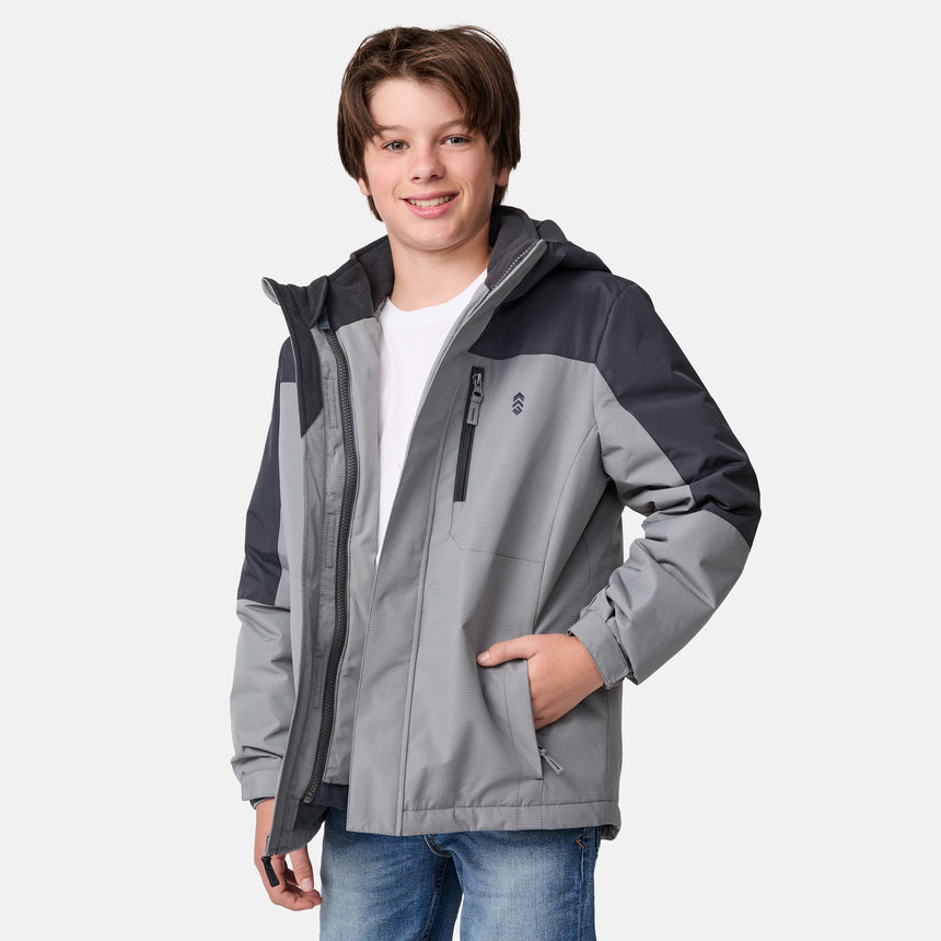 [River is wearing size M.] Young boy wearing the Free Country Boys' 3-in-1 Systems Jacket in a light grey with a hood and subtle chest pattern, smiling against a plain background.