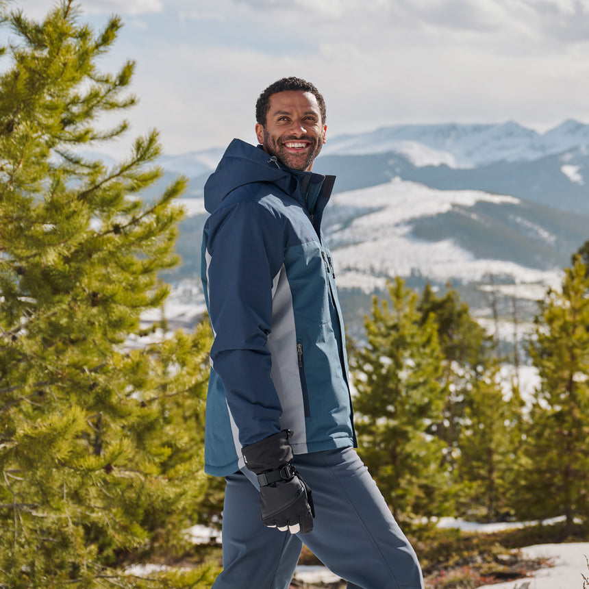Man in a blue jacket standing in a snowy landscape with mountains and trees.