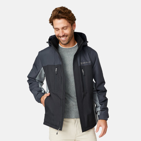 [Matt is 6'2" wearing size M.] Men’s Free Country black zip-front jacket with gray hood, sleeves, and side panels—features dual zippered chest pockets and side pockets for practical, cold-weather functionality. Paired with beige pants for a clean, transitional outdoor look.