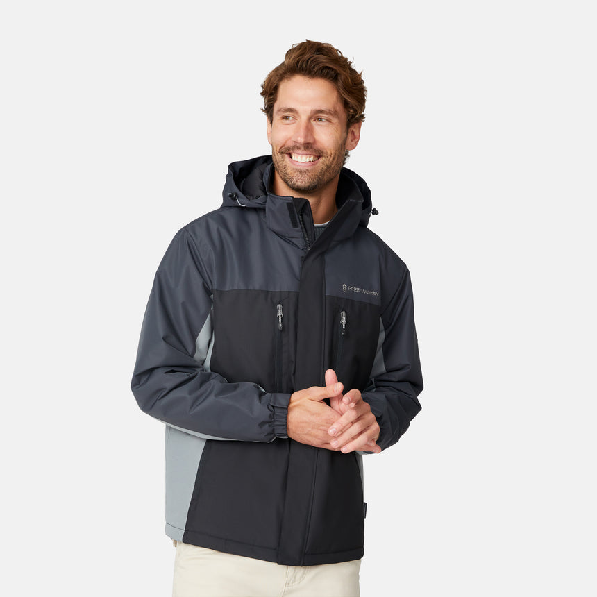 [Matt is 6'2" wearing size M.] Men’s Free Country black zip-front jacket with gray hood, sleeves, and side panels—features dual zippered chest pockets and side pockets for practical, cold-weather functionality. Paired with beige pants for a clean, transitional outdoor look.