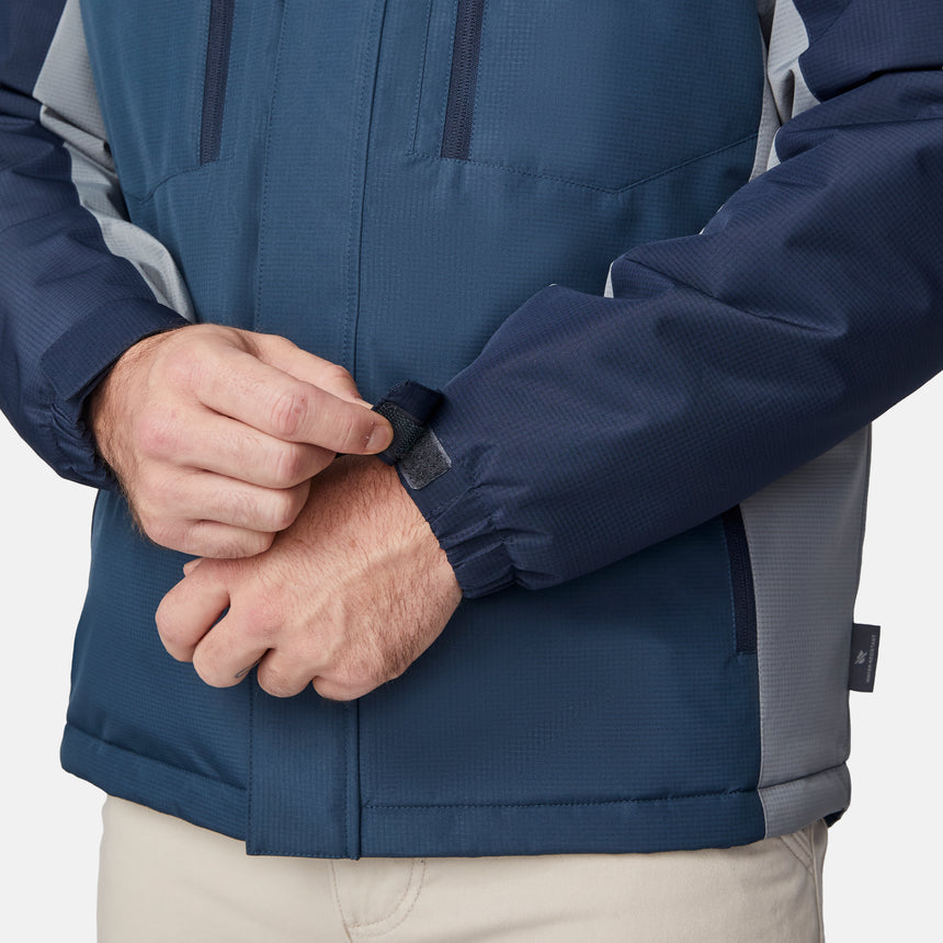 [Joe is 6'0" wearing size M.] Men’s Free Country hooded zip-front jacket in blue with gray paneling—featuring dual vertical zippered chest pockets and side hand pockets. Paired with light khaki pants for a clean, functional look suited to cold-weather layering and outdoor versatility.