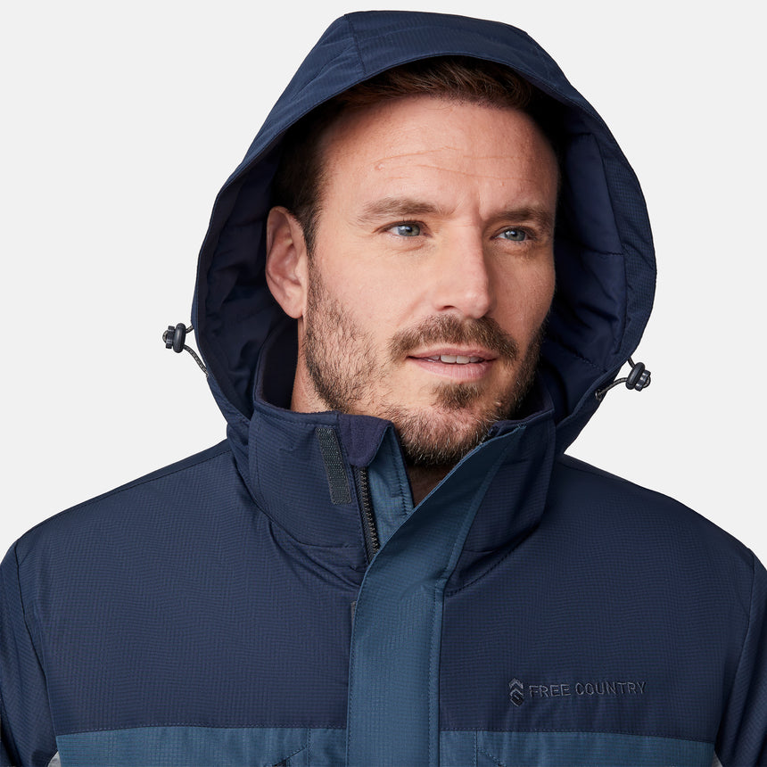 [Joe is 6'0" wearing size M.] Men’s Free Country hooded zip-front jacket in blue with gray paneling—featuring dual vertical zippered chest pockets and side hand pockets. Paired with light khaki pants for a clean, functional look suited to cold-weather layering and outdoor versatility.