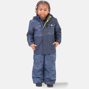 A young child with braided hair stands facing forward, wearing the Free Country Boys' Toddler Boarder Jacket and Bib Pant Set in blue pattern, hands in pockets, styled with black shoes against a plain white background.