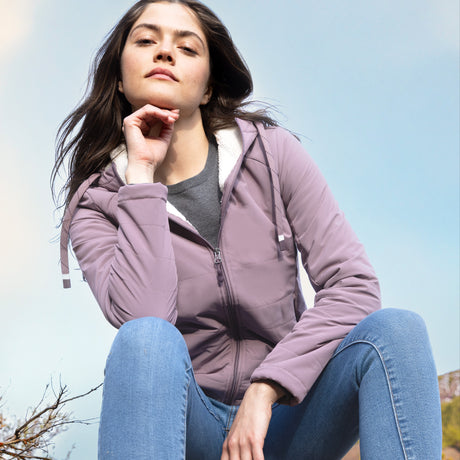 A woman with long brown hair wears the Free Country Women's AirLoft Quilted Jacket in mauve and blue jeans as she sits outdoors, confidently resting her chin on her hand against a pale blue sky.