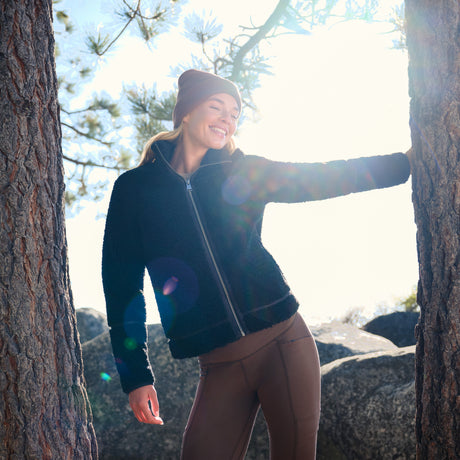 A woman wearing a black fleece zip-up jacket and brown leggings stands outdoors between two tree trunks in bright sunlight, smiling and touching the tree on her right.