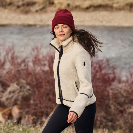 Woman wearing a cream jacket and red beanie standing by a body of water with autumn foliage.