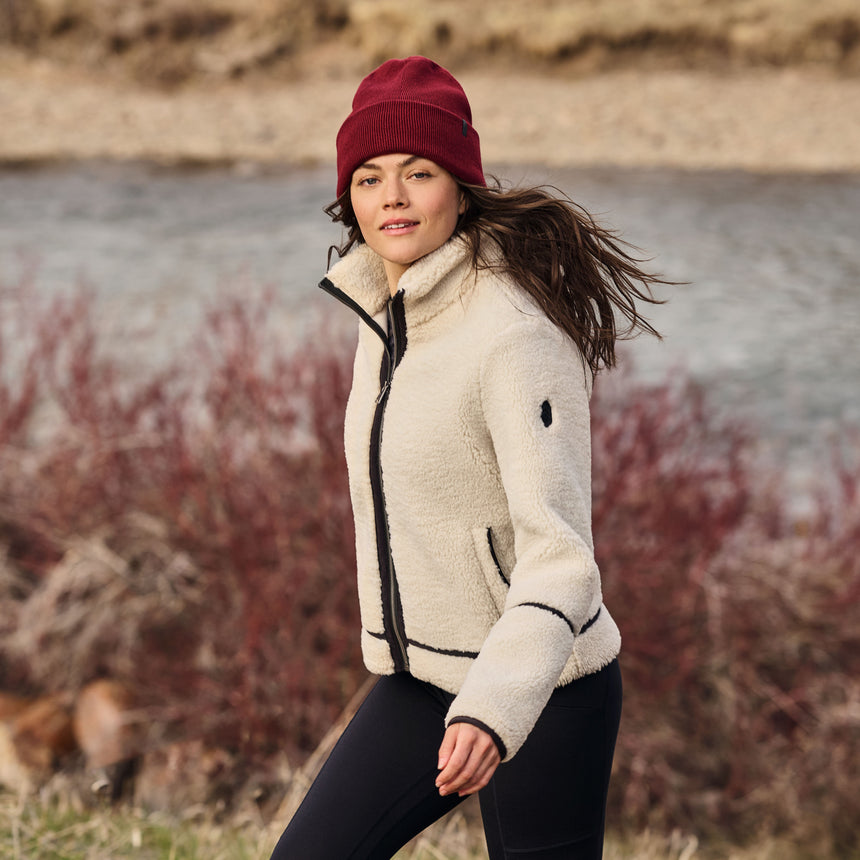 Woman wearing a cream jacket and red beanie standing by a body of water with autumn foliage.