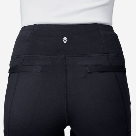 A person wearing Free Country Women's Trail 2 Town Flare Pants in black, featuring a small white chevron logo on the waistband and two horizontal back pocket details, viewed from the back.