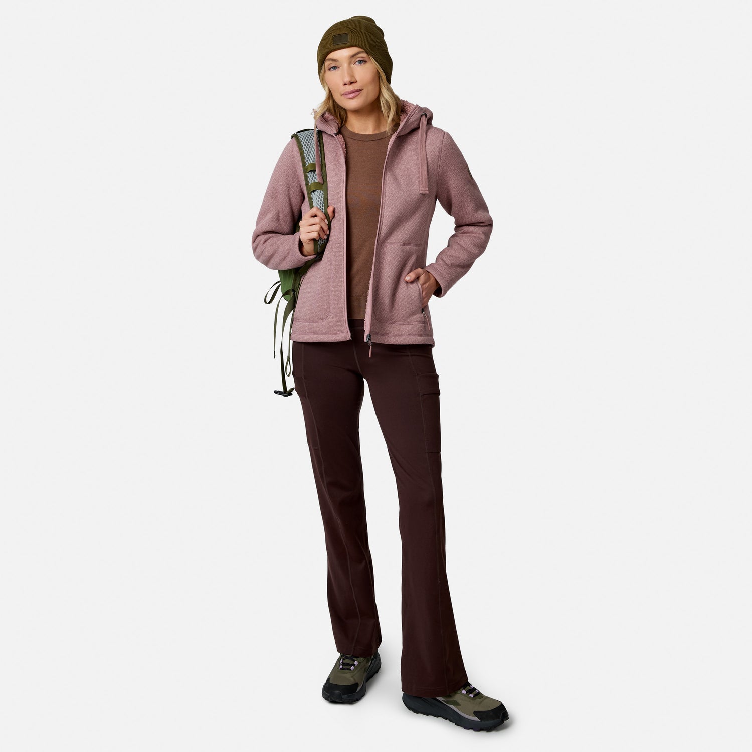 A woman in outdoor wear, featuring Free Country Women’s Trail 2 Town Flare Pants, a pink jacket, brown shirt, boots, and green beanie, stands holding climbing rope over her shoulder against a plain white background.