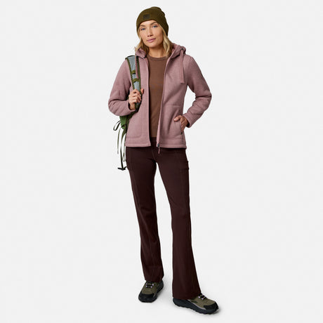 A woman in outdoor wear, featuring Free Country Women’s Trail 2 Town Flare Pants, a pink jacket, brown shirt, boots, and green beanie, stands holding climbing rope over her shoulder against a plain white background.