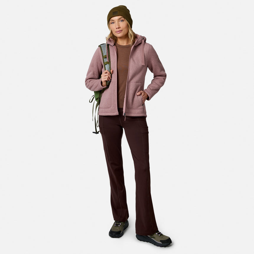 A woman in outdoor wear, featuring Free Country Women’s Trail 2 Town Flare Pants, a pink jacket, brown shirt, boots, and green beanie, stands holding climbing rope over her shoulder against a plain white background.