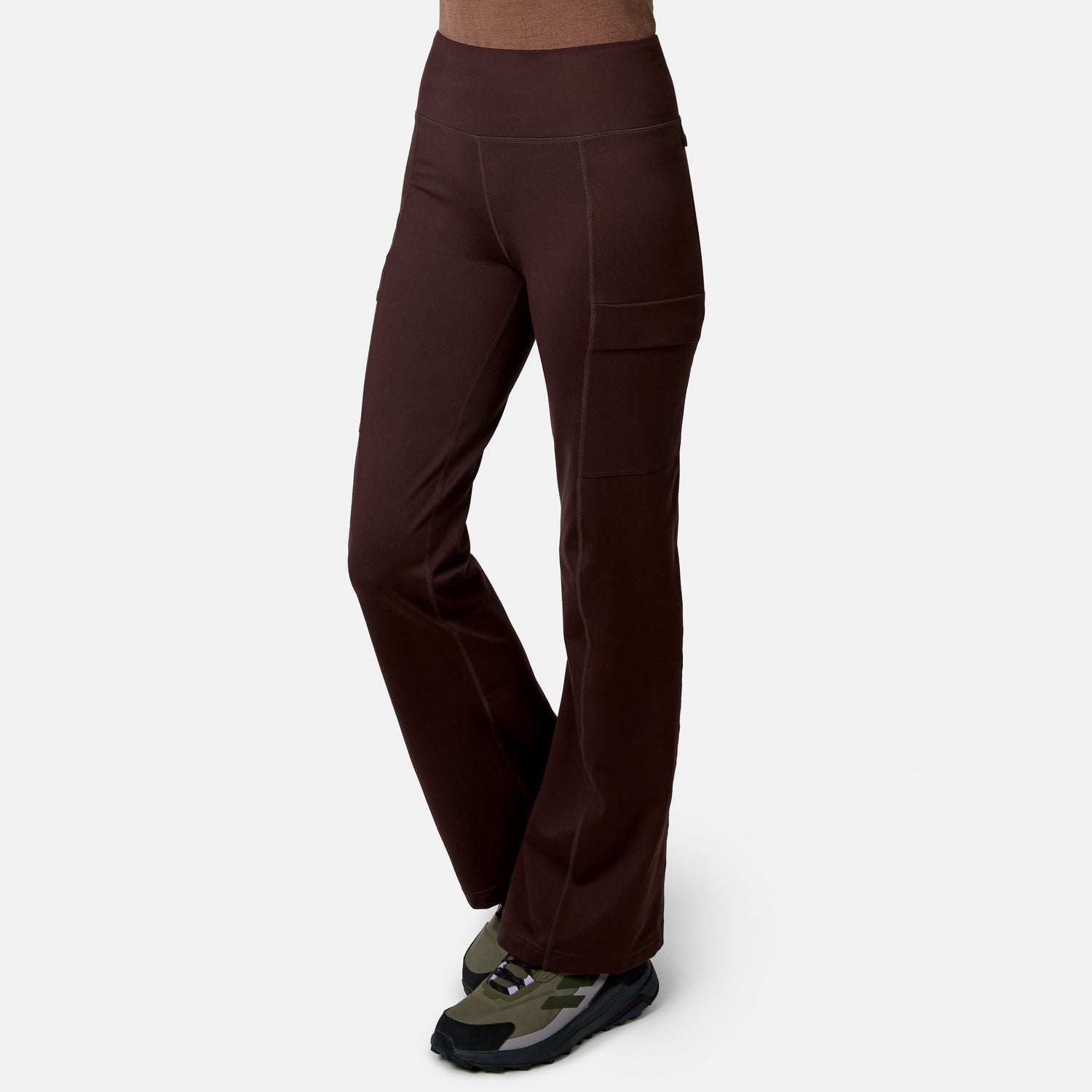 Model wears Free Country Women’s Trail 2 Town Flare Pants with visible stitching and side pockets, paired with green and black athletic shoes, against a white background.