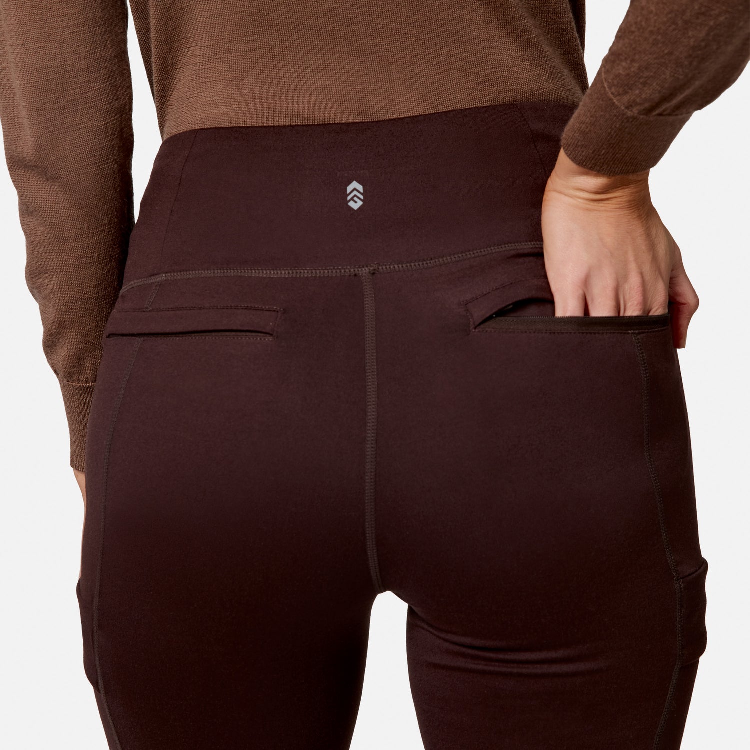 Person wears Free Country Women’s Trail 2 Town Flare Pants in dark brown and a brown top, shown from the back with their right hand partially inside the right back pocket. Background is plain white.