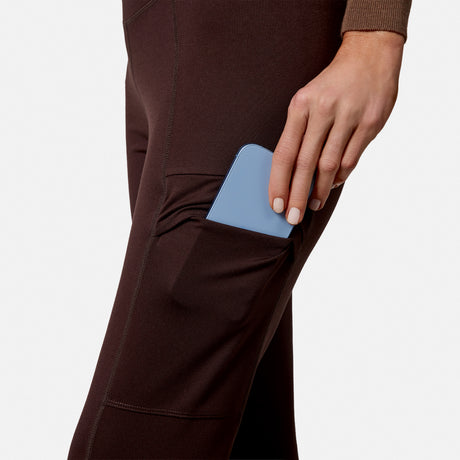 A person in Free Country Women’s Trail 2 Town Flare Pants (UPF 50+) slips a light blue phone into the side pocket. Only their hand, phone, and lower torso appear against a plain white background.