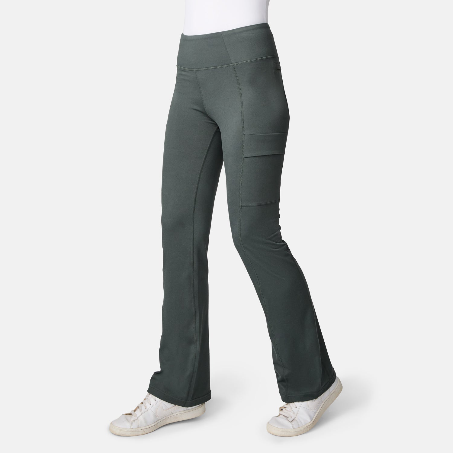 A person wears Free Country Women’s Trail 2 Town Flare Pants in Loden with side pockets and white sneakers, standing against a plain white background.