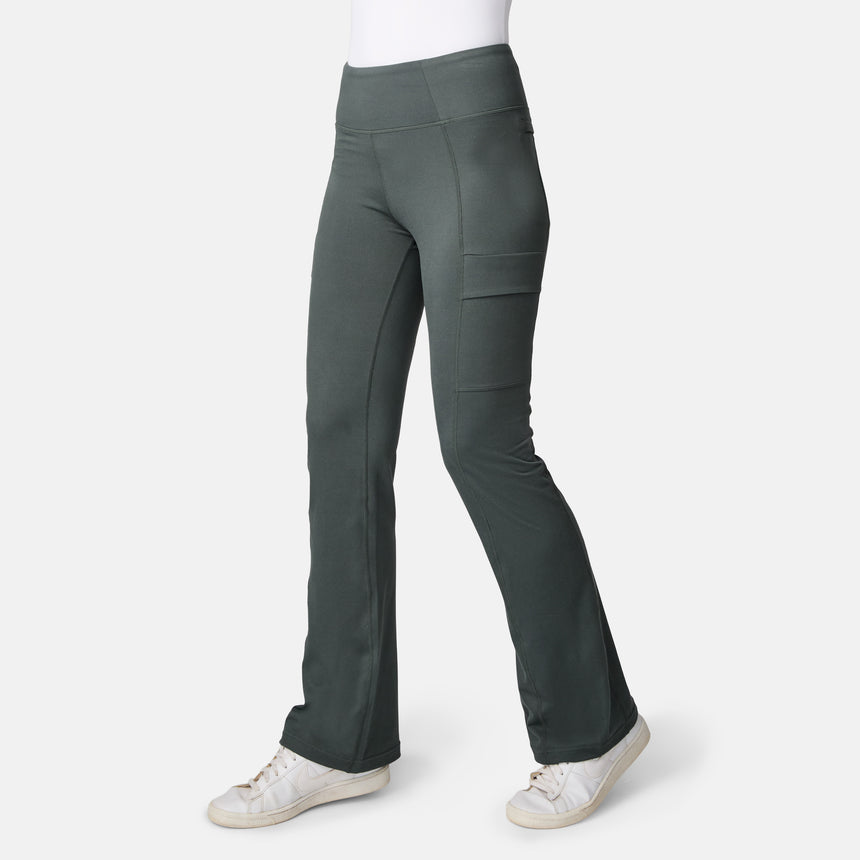 A person wears Free Country Women’s Trail 2 Town Flare Pants in Loden with side pockets and white sneakers, standing against a plain white background.