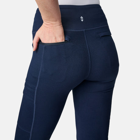 A woman models Free Country Women’s Trail 2 Town Flare Pants in navy, one hand in the back pocket, showing off the waistband and stitching. The simple, light background highlights these stylish flare pants.