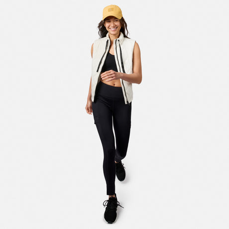 A smiling woman wearing a yellow baseball cap, white vest, black sports bra, and Free Country Women’s Trail 2 Town Legging walks forward.