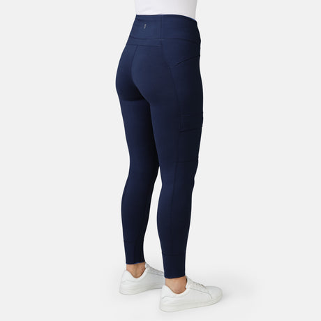 A woman is shown from the back wearing Free Country Women's Trail 2 Town Legging in navy blue, featuring a high rise waistband and side pockets, paired with a white top and sneakers, against a plain light background.