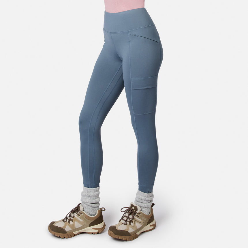Free Country Women’s Trail 2 Town Legging in blue, featuring a high waist, four-way stretch, a zippered hip pocket, and a side cargo pocket, worn with gray socks and tan hiking sneakers.