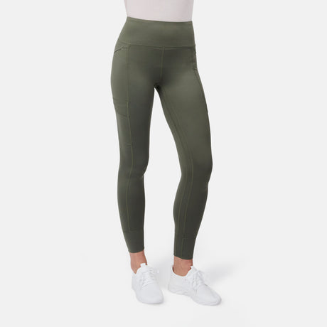 A woman is shown from the waist down wearing Free Country Women's Trail 2 Town Legging in olive green with a high rise waistband and side pockets, paired with white sneakers and a white top against a plain white background.