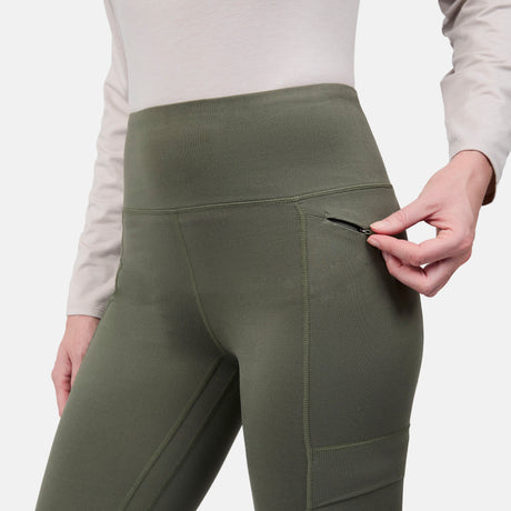 A person wearing Free Country Women's Trail 2 Town Legging in green with a high-rise waistband is unzipping a side pocket. Only the lower torso and hand are visible.