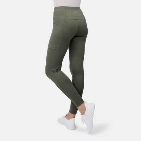 A person wears Free Country Women's Trail 2 Town Legging in olive green, featuring a high-rise waistband and water-repellent fabric, paired with white sneakers, standing with one leg bent and hands in the pockets against a white background.