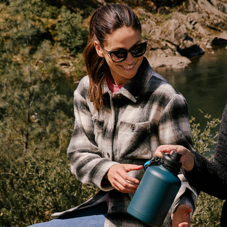 A woman in the Clearance Women's Hayley Plaid Shacket smiles by a riverside, reaching for a blue water bottle as sunlight filters through trees and rocks in the background. Sunglasses rest on her face, completing her outdoor look.