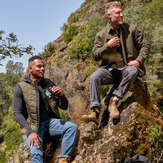 Man wearing the Free Country Men's FreeCycle® Stimson Puffer Vest in green, featuring quilted design, REPREVE® poly fill, and a vertical zip, layered over a black long-sleeve shirt with dark jeans.