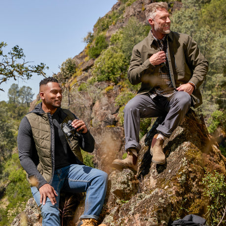 Man wearing the Free Country Men's FreeCycle® Stimson Puffer Vest in green, featuring quilted design, REPREVE® poly fill, and a vertical zip, layered over a black long-sleeve shirt with dark jeans.